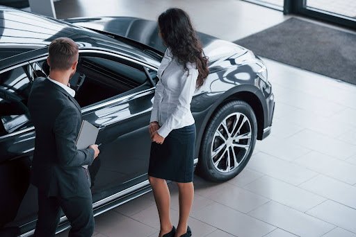 Bearded salesman with clipboard speaks to woman customer by dark gray Luxury car rental in modern dealership showroom.
